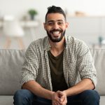 Portrait of handsome young Arab man smiling and looking at camera, sitting on sofa at home